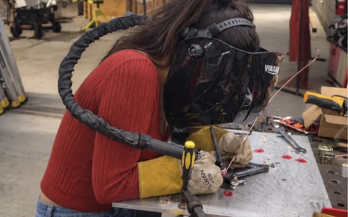Victoria TIG welding at a shop welding table, wearing a welding helmet and gloves, torch in one hand and filler rod in the other