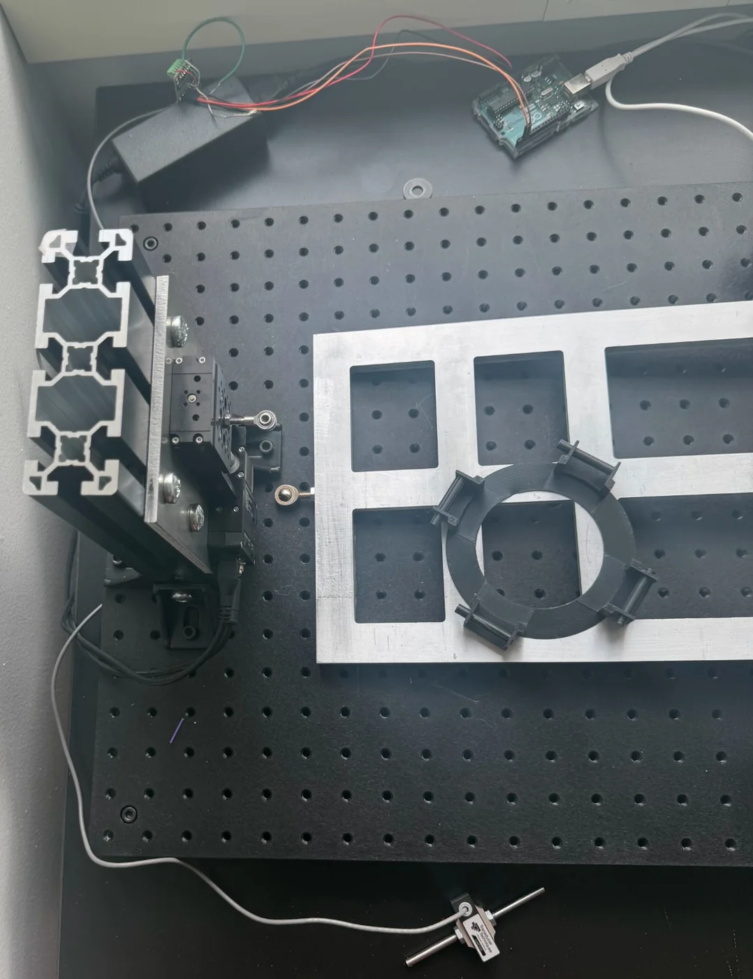 Top view of the rig on the lab bench: the extrusion column with the Zaber stage and rod end on the left, the machined aluminum lid with a black printed clamp ring over a black optical breadboard, an Arduino Uno at the top, and the load cell on its rod lying unmounted at the bottom